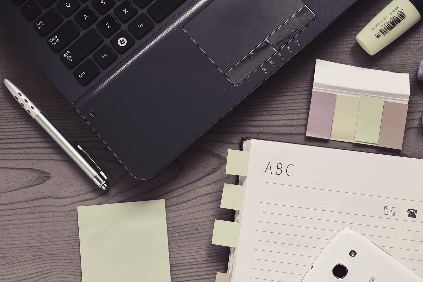 A laptop, notebook, sticky notes and pen on a wooden desk